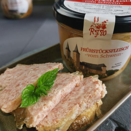 hausgemachtes Frühsücksfleisch vom Schwein in Scheiben geschnitten, angerichtt auf einer Scheibe Brot, garniert mit frischer Petersilie neben Glaskonserve