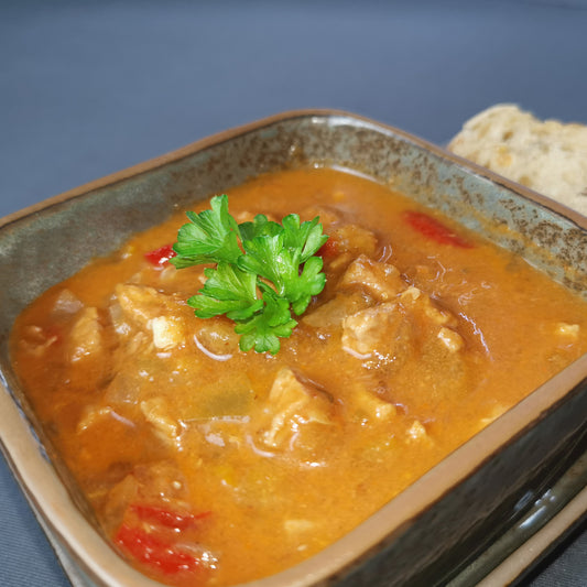 hausgemachter Schweinegulasch angerichtet in einer viereckigen Schale neben einer scheibe Brot garnier tmit frischer Petersilie